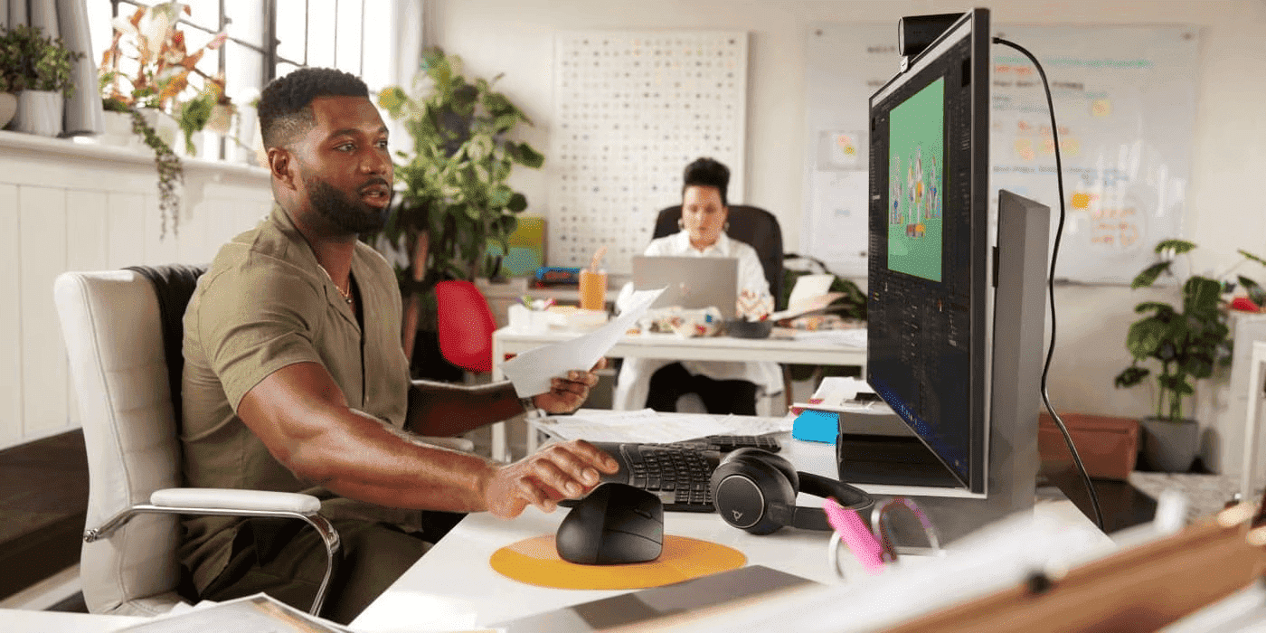 A man in an olive shirt reviews documents while working at a dual-monitor setup reaching for the HP 920 Ergonomic Vertical Mouse, with a colleague working at a laptop in the background of a modern office.