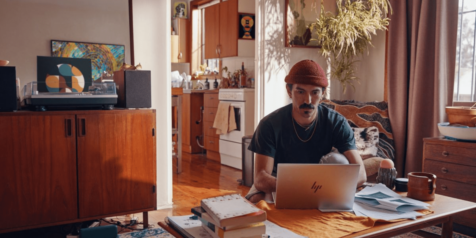 A man with a mustache and red beanie works on a laptop at a cluttered coffee table in a cozy apartment, surrounded by vintage furniture and eclectic decor.