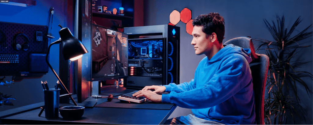A person in a blue hoodie sits at a gaming setup with an open PC case showing internal components, illuminated by blue and red lighting in a darkened room with hexagonal wall decorations
