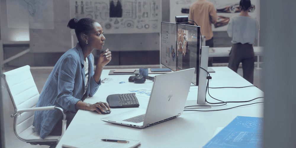 A woman working thoughtfully at a laptop connecteed to an external monitor in a modern office with colleagues collaborating on design projects in the background